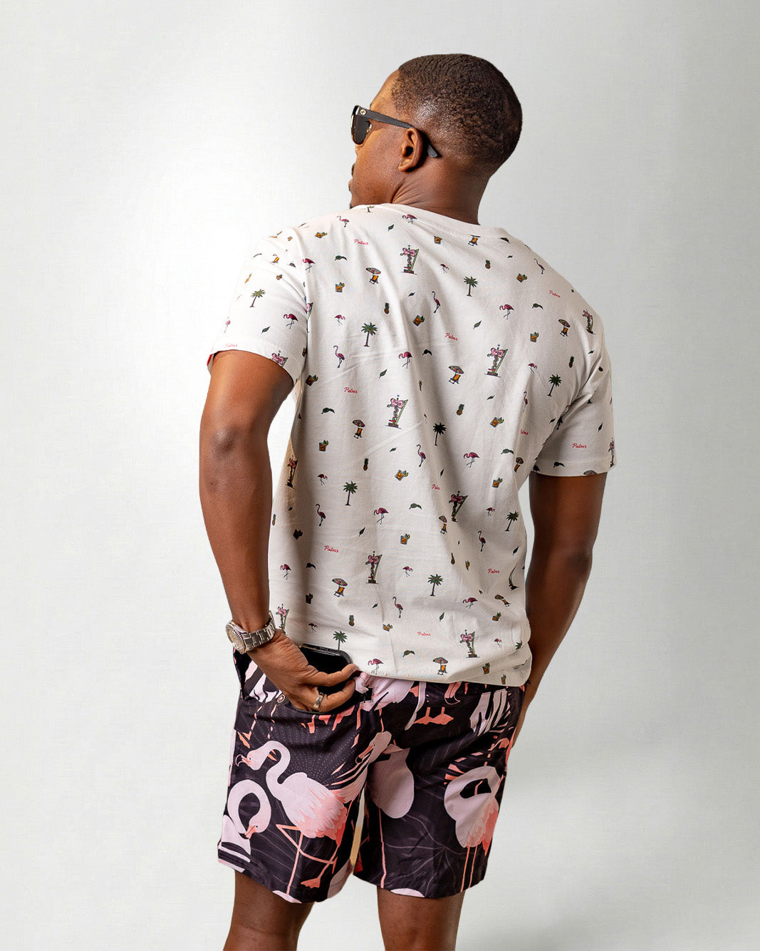 Person wearing a white shirt with a palm tree pattern and black shorts with pink flamingo pattern on a plain background