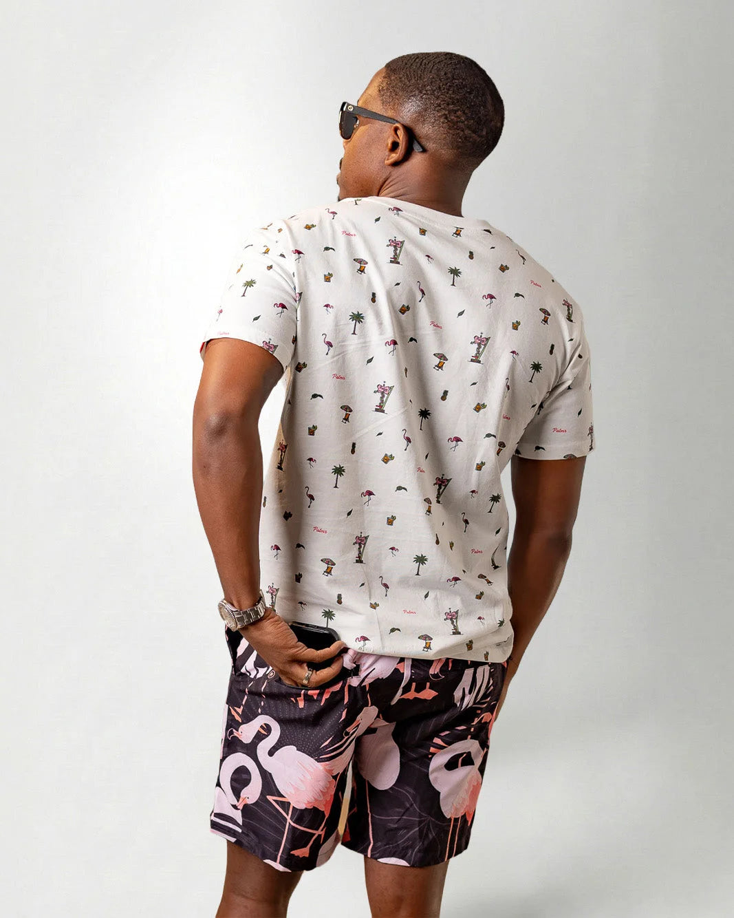 Person wearing a white shirt with a palm tree pattern and black shorts with pink flamingo pattern on a plain background
