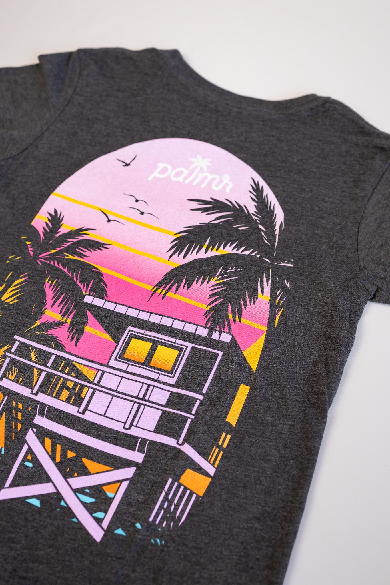 Gray t-shirt with a colorful beach house and palm tree design on a white background