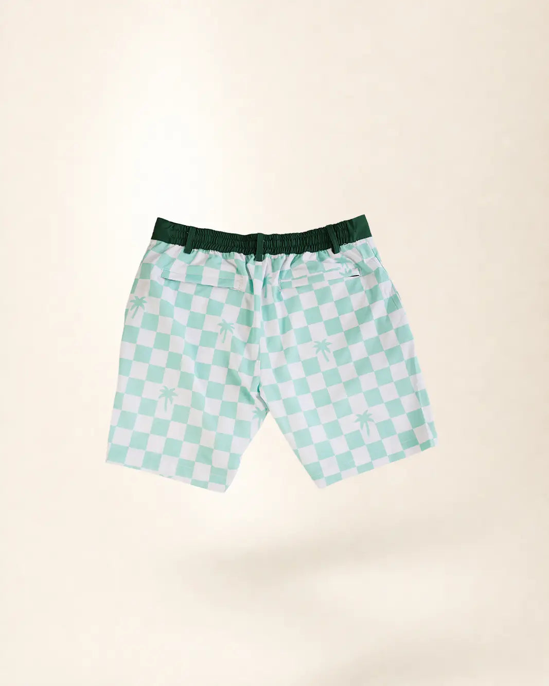 A pair of men's swim trunks featuring a mint green and white checkered pattern with small palm tree motifs, a dark green elastic waistband, and a white drawstring, shown against a solid cream background.