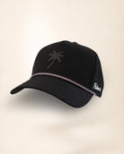 A black trucker-style baseball cap with a black palm tree logo on the front, a silver chain detail across the brim, and white script embroidery on the side mesh, shown against a plain off-white background.