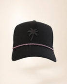A black baseball cap featuring a tonal black palm tree logo on the front and a thin pink chain detail running horizontally around the crown, set against a solid light beige background.