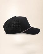 A black baseball cap with perforated side panels and a curved brim, featuring a thin pink and white patterned trim along the edge of the brim, set against a solid light beige background.