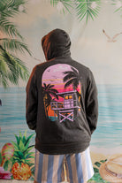 Person wearing a hoodie with a palm tree and beach house design, standing in front of a tropical-themed wall.