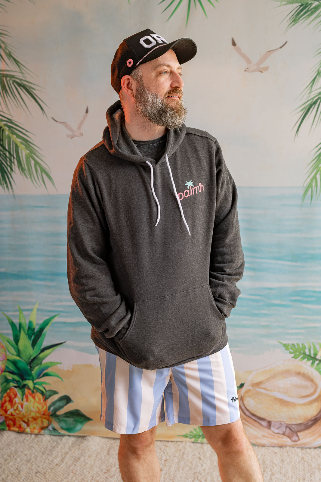 Man wearing a dark hoodie with a logo and striped shorts standing in front of a beach-themed wall mural.