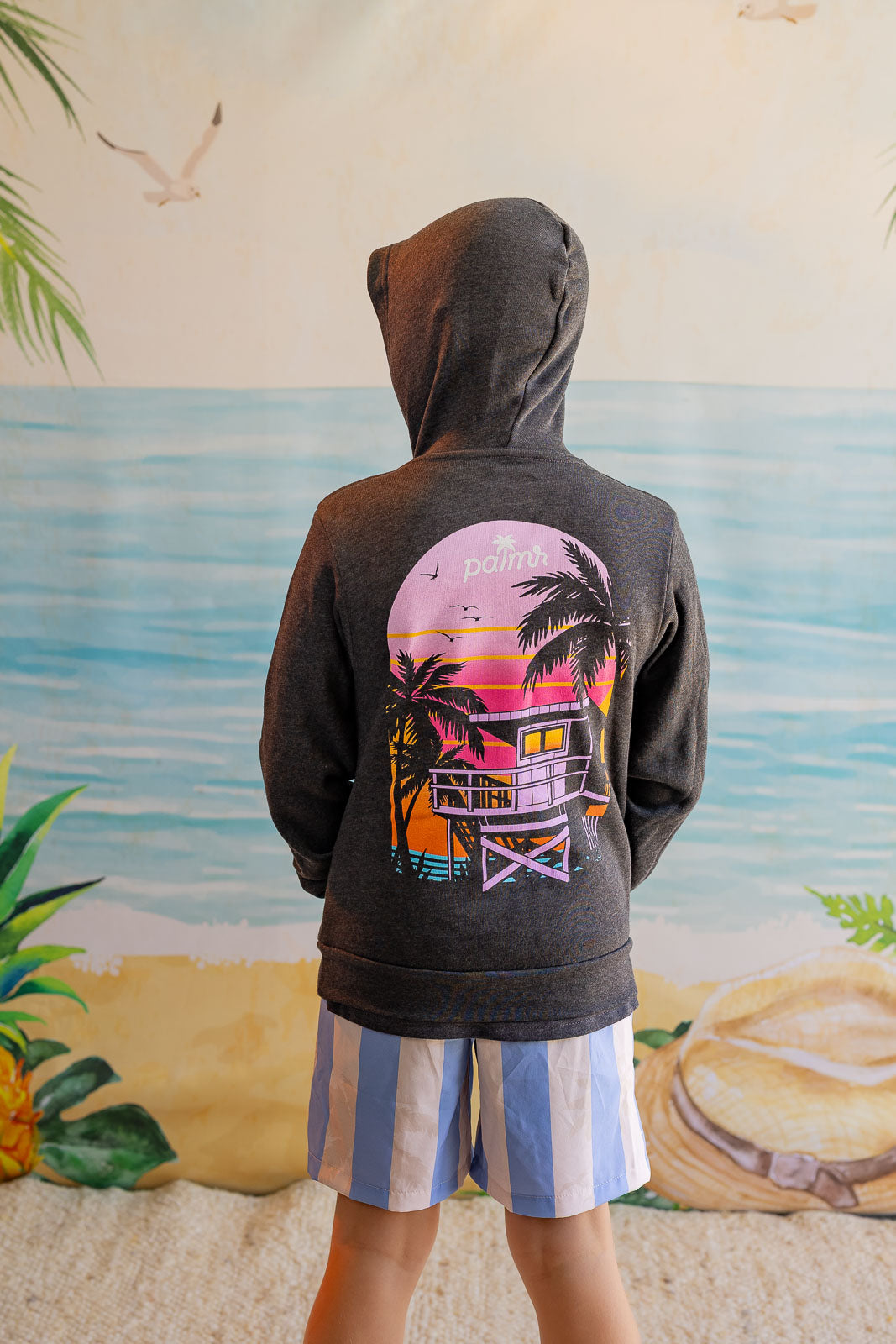 Person wearing a hoodie with a colorful lifeguard tower design in front of a beach mural.
