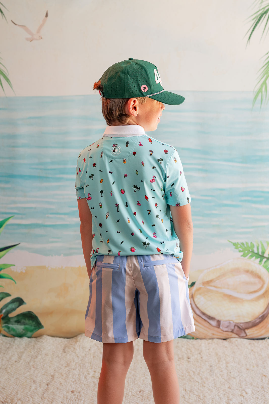 Child wearing a patterned shirt and cap, standing against a beach-themed wall.