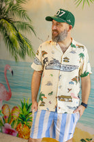 Man wearing a tropical-themed shirt and cap with a colorful background featuring flamingos and palm trees.