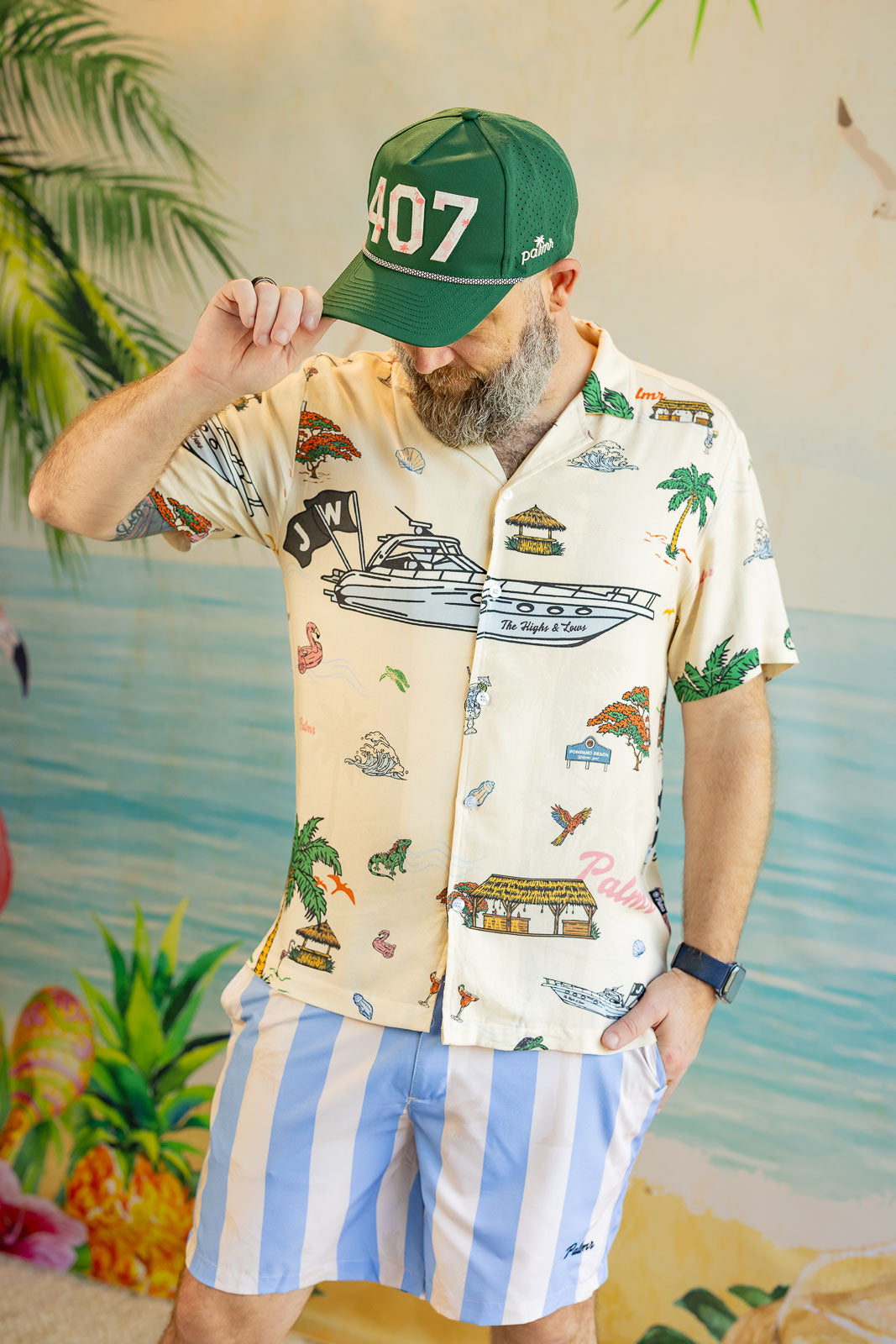 Man wearing a patterned shirt and striped shorts with a green cap, standing against a tropical-themed background.