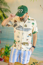 Man wearing a patterned shirt and striped shorts with a green cap, standing against a tropical-themed background.