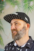 Man wearing a black cap with white patterns and text, standing against a blurred green leafy background.