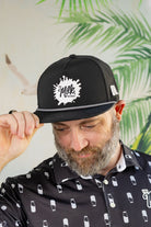 Man wearing a black cap with a logo and a patterned shirt against a leafy background