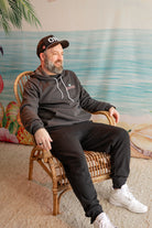 Man sitting on a wicker chair wearing a hoodie with a logo, in a room with a decorative wall.
