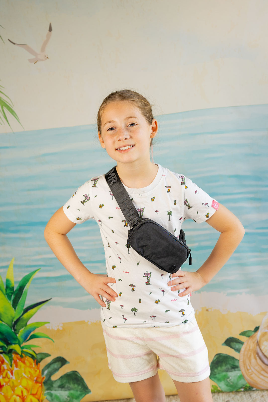 Child wearing a patterned shirt and shorts with a black waist bag, standing in front of a beach-themed wall.