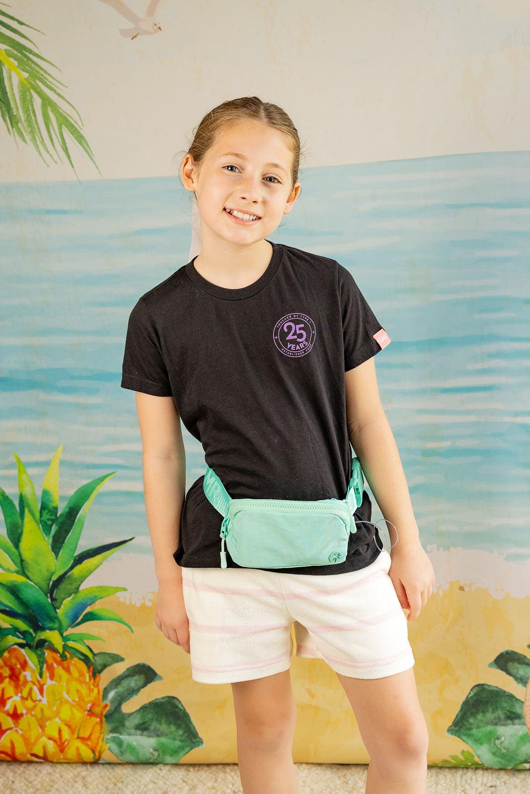 Child wearing a black t-shirt with a logo and white shorts, standing in front of a colorful mural.