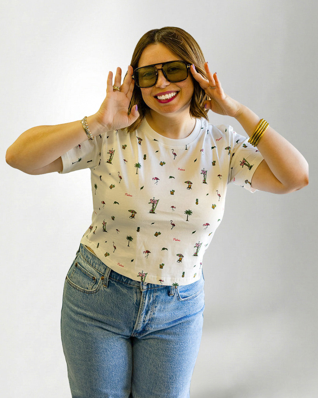 Woman wearing a patterned shirt and jeans with sunglasses on a plain background