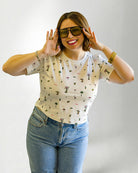 Woman wearing a patterned shirt and jeans with sunglasses on a plain background