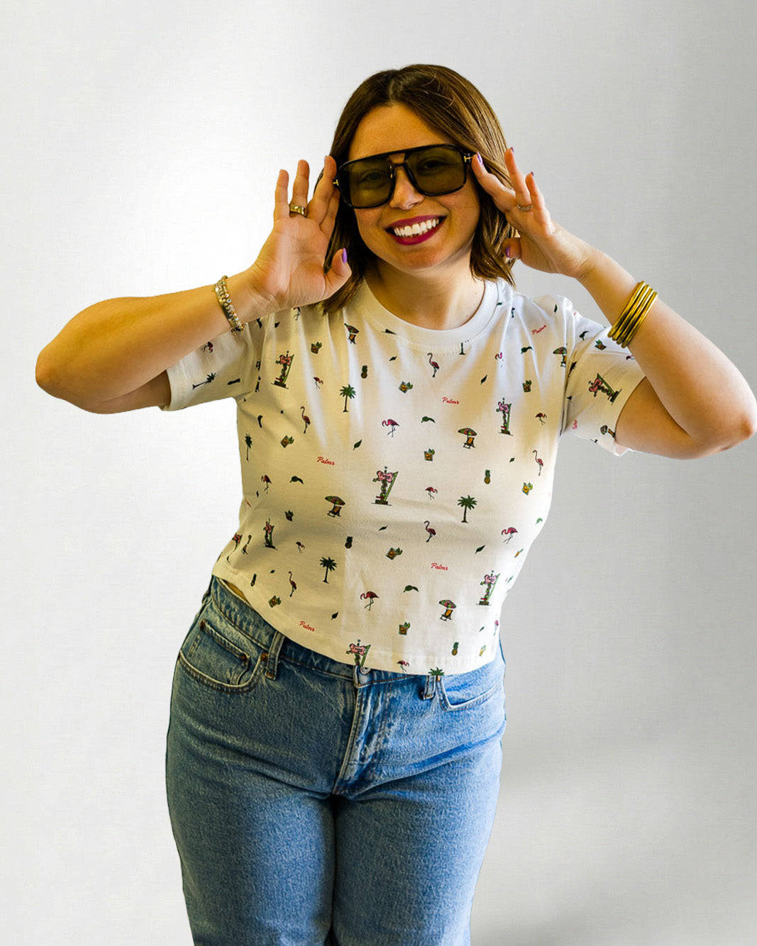 Woman wearing a patterned shirt and jeans with sunglasses on a plain background
