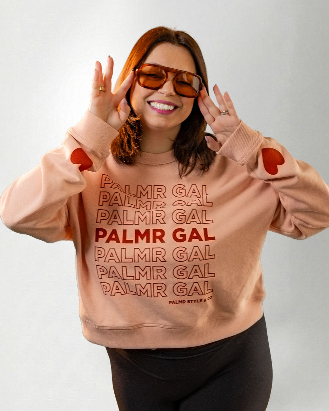 Person wearing a peach sweatshirt with 'PALMR GAL' text and heart designs on a plain background