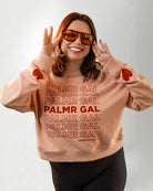 Person wearing a peach sweatshirt with 'PALMR GAL' text and heart designs on a plain background