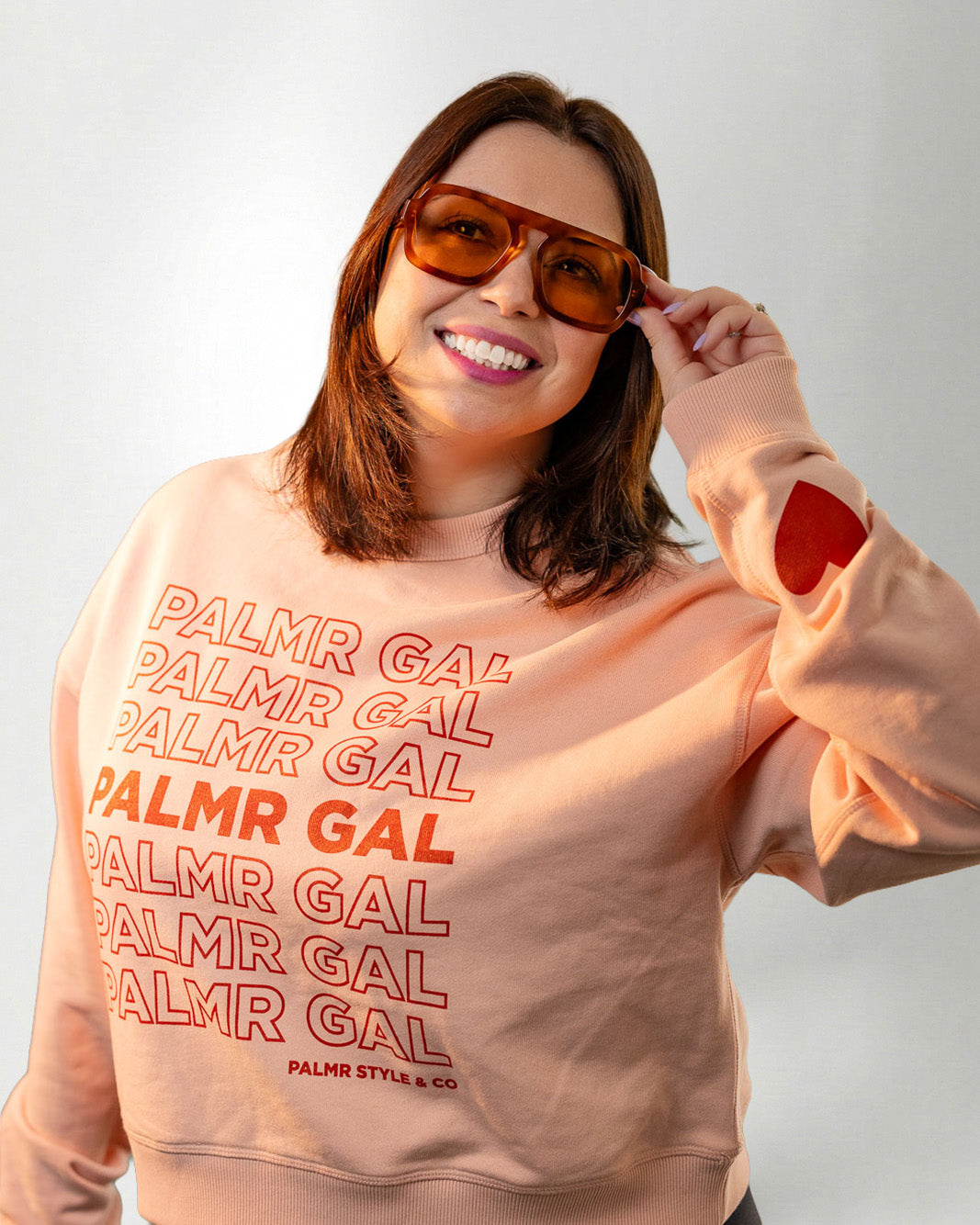 Person wearing a peach sweatshirt with 'PALMR GAL' text and heart designs on a plain background