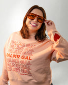 Person wearing a peach sweatshirt with 'PALMR GAL' text and heart designs on a plain background