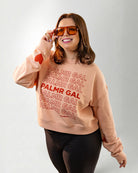 Person wearing a peach sweatshirt with 'PALMR GAL' text and heart designs on a plain background