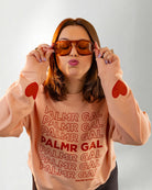 Person wearing a peach sweatshirt with 'PALMR GAL' text and heart designs on a plain background