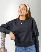 Woman wearing a navy blue sweatshirt with 'PALMR' branding, holding a floral-patterned bag against a white background.