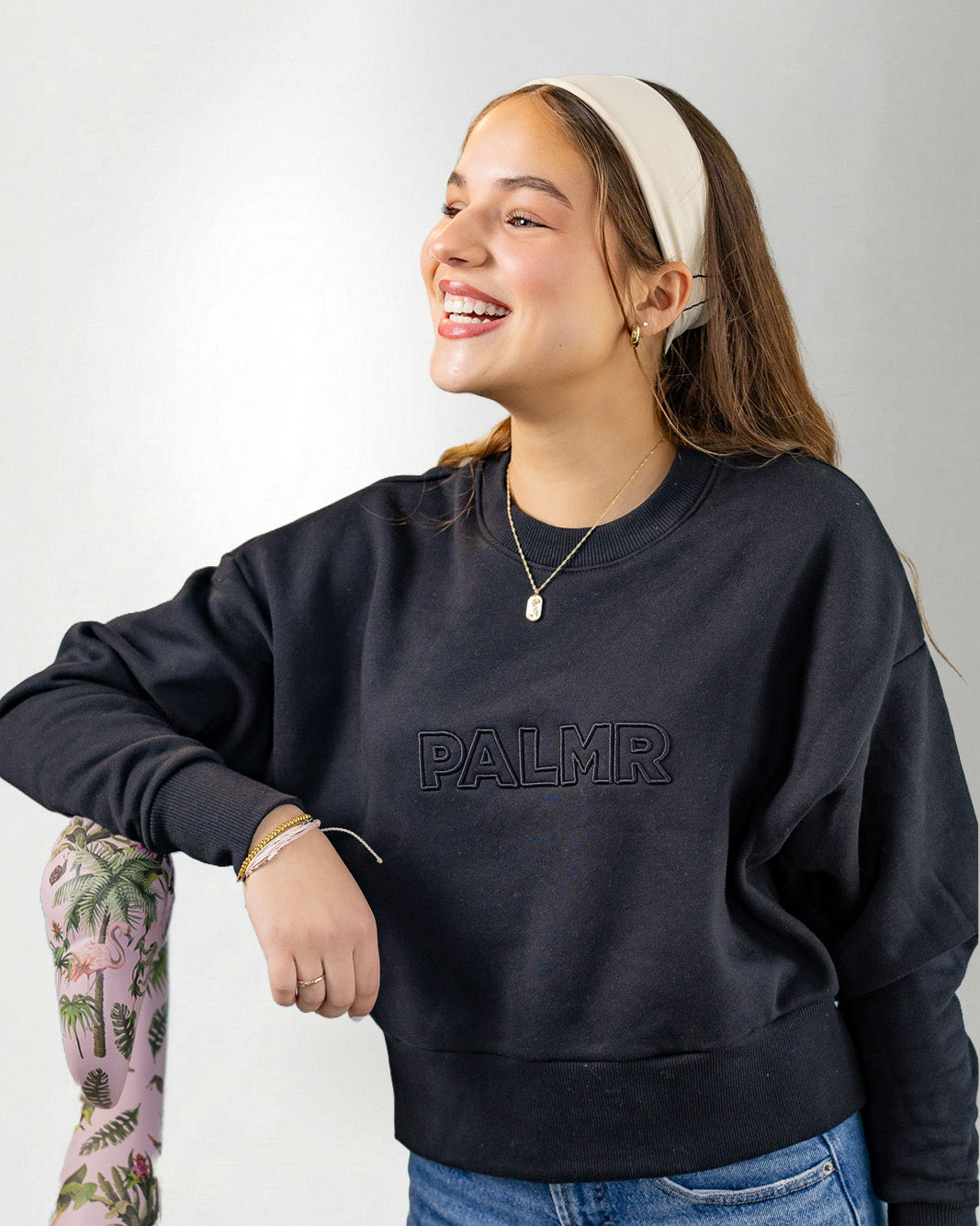 Woman wearing a navy blue sweatshirt with 'PALMR' branding, holding a floral-patterned bag against a white background.