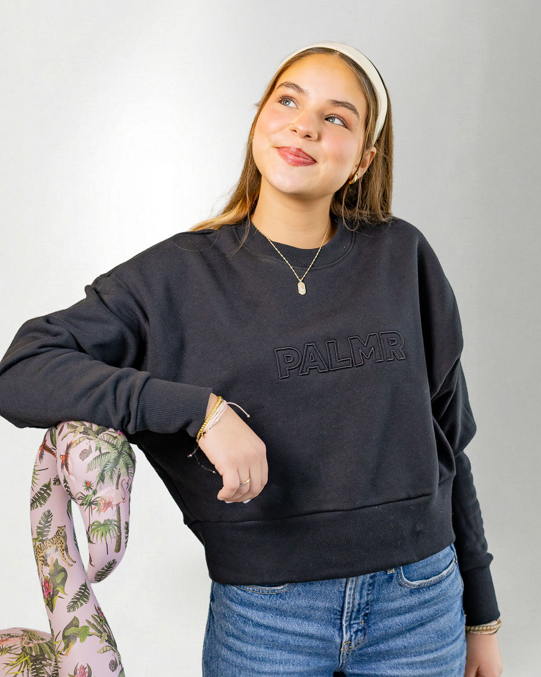 Woman wearing a navy blue sweatshirt with 'PALMR' branding, holding a floral-patterned bag against a white background.