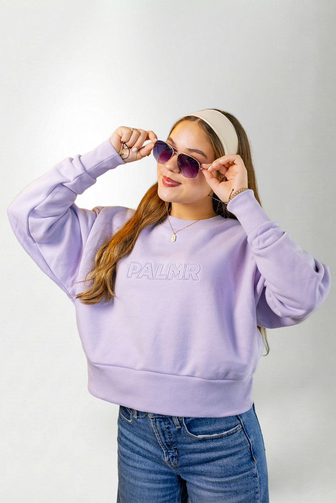Woman wearing a lavender sweatshirt with 'PALM' branding, sunglasses, and a headband on a white background.