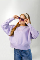 Woman wearing a lavender sweatshirt with 'PALM' branding, sunglasses, and a headband on a white background.