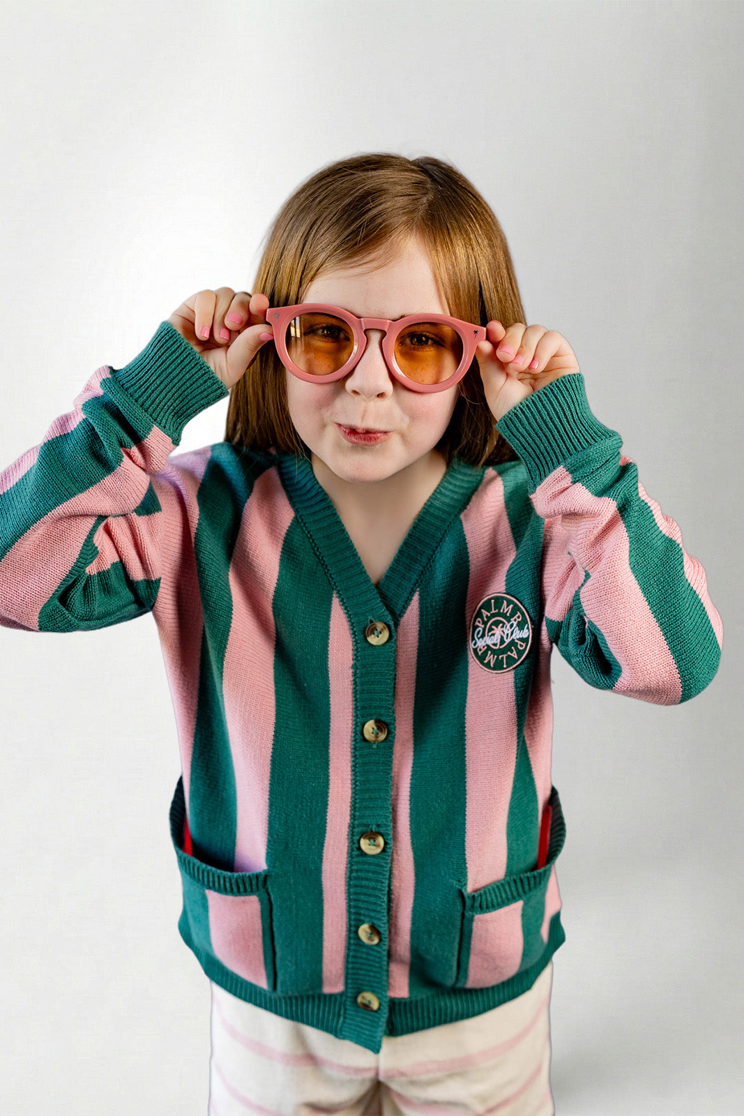 Child wearing a pink and green striped cardigan with a logo, holding pink sunglasses.