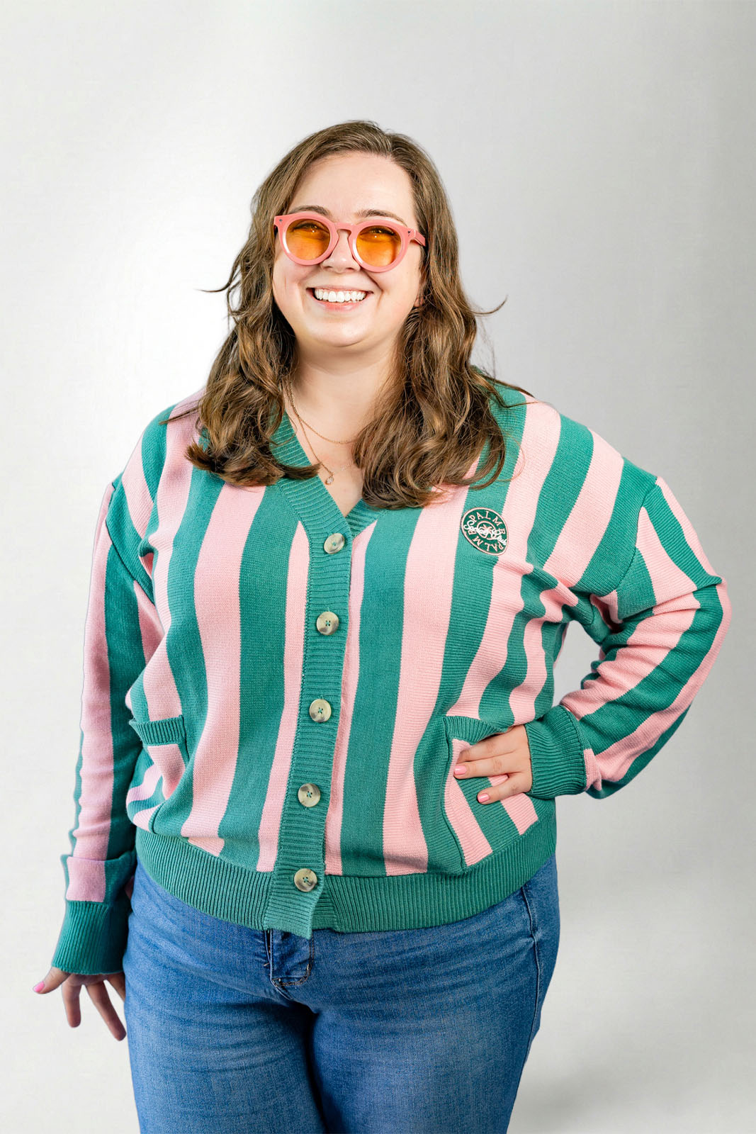 Woman wearing a green and pink striped cardigan with a logo, smiling against a plain background
