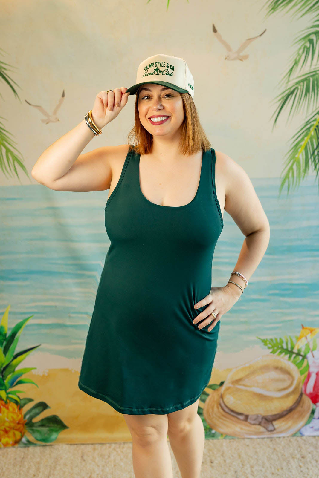 Woman wearing a green dress and white cap with palm tree design, standing in front of a tropical-themed backdrop.