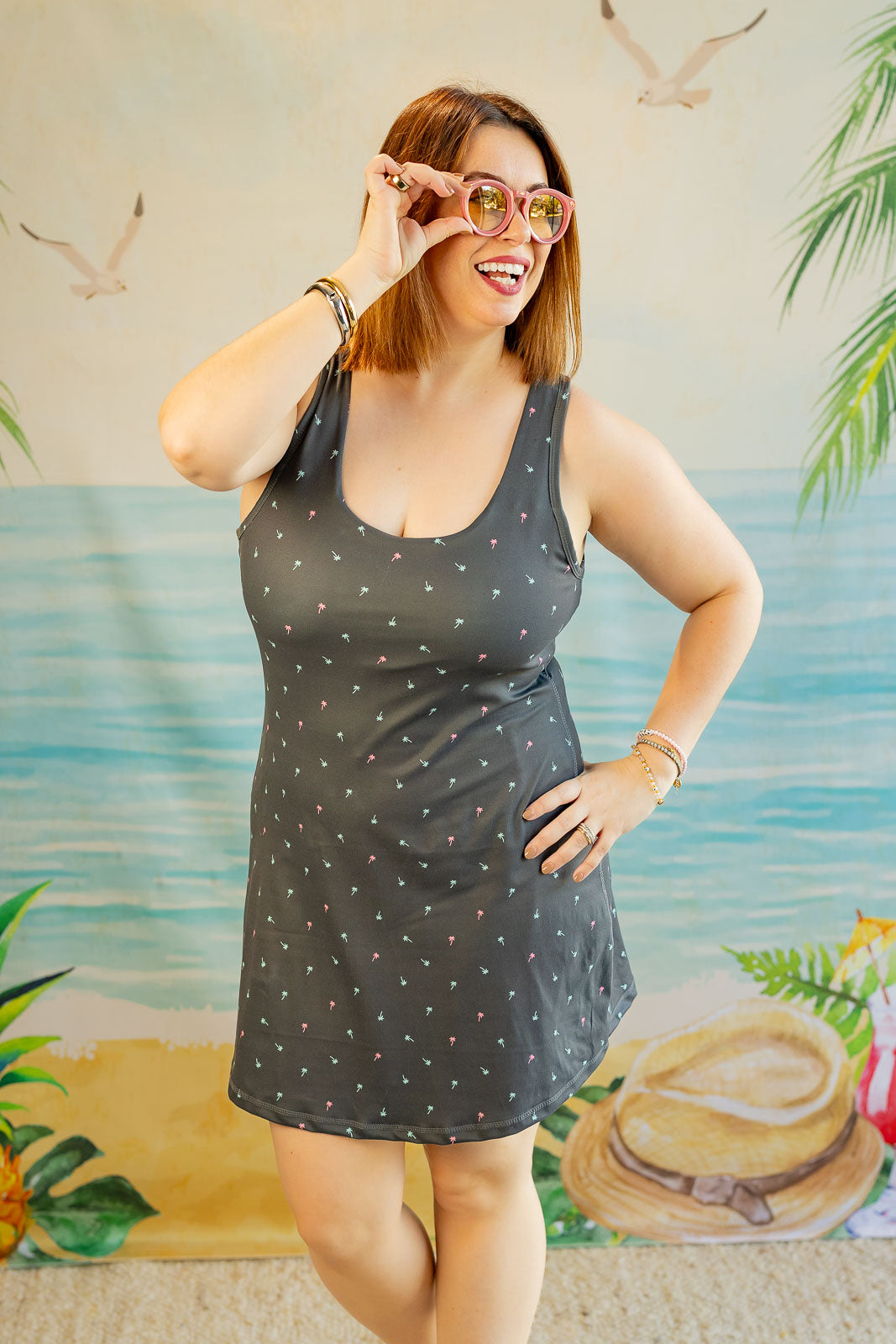 Woman wearing a dark gray tank dress with white polka dots in front of a tropical-themed backdrop.