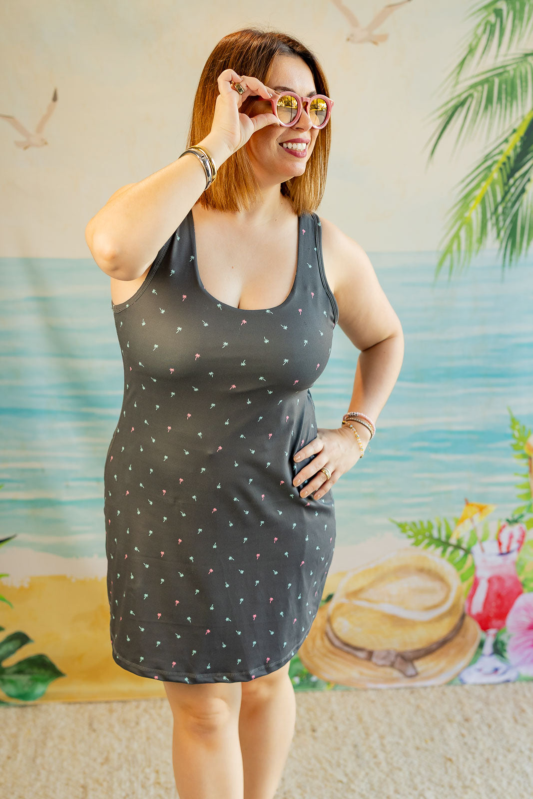 Woman wearing a dark gray sleeveless dress with white polka dots, standing in front of a tropical-themed wall.