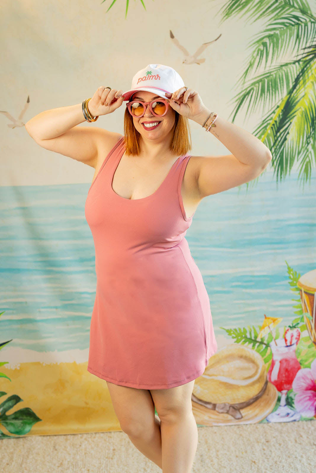 Woman in a pink dress and white cap posing in front of a tropical-themed backdrop