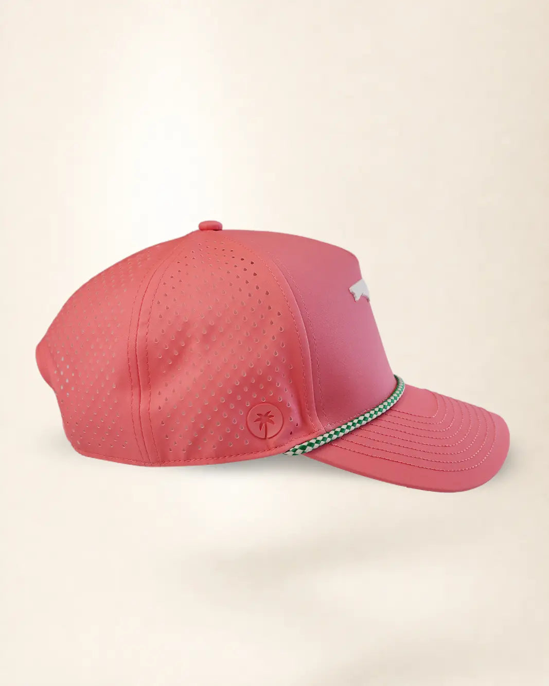 A coral pink performance baseball cap with perforated side panels, a small white logo on the side, and a green and white checkered trim detail on the brim, shown against a plain cream background.