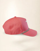 A coral pink performance baseball cap with perforated side panels, a small white logo on the side, and a green and white checkered trim detail on the brim, shown against a plain cream background.