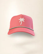 A pink baseball cap featuring a white palm tree logo on the front and a decorative green and white beaded trim along the brim, set against a plain off-white background.