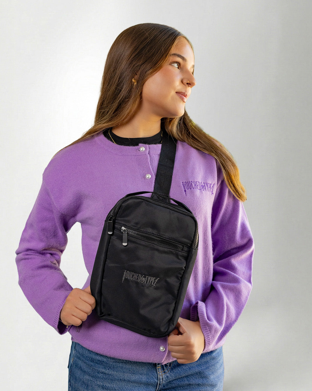 Girl using a "Touched by Type one purple cardigan" and a "Touched by type 1" Black crossbody bag against a gray background