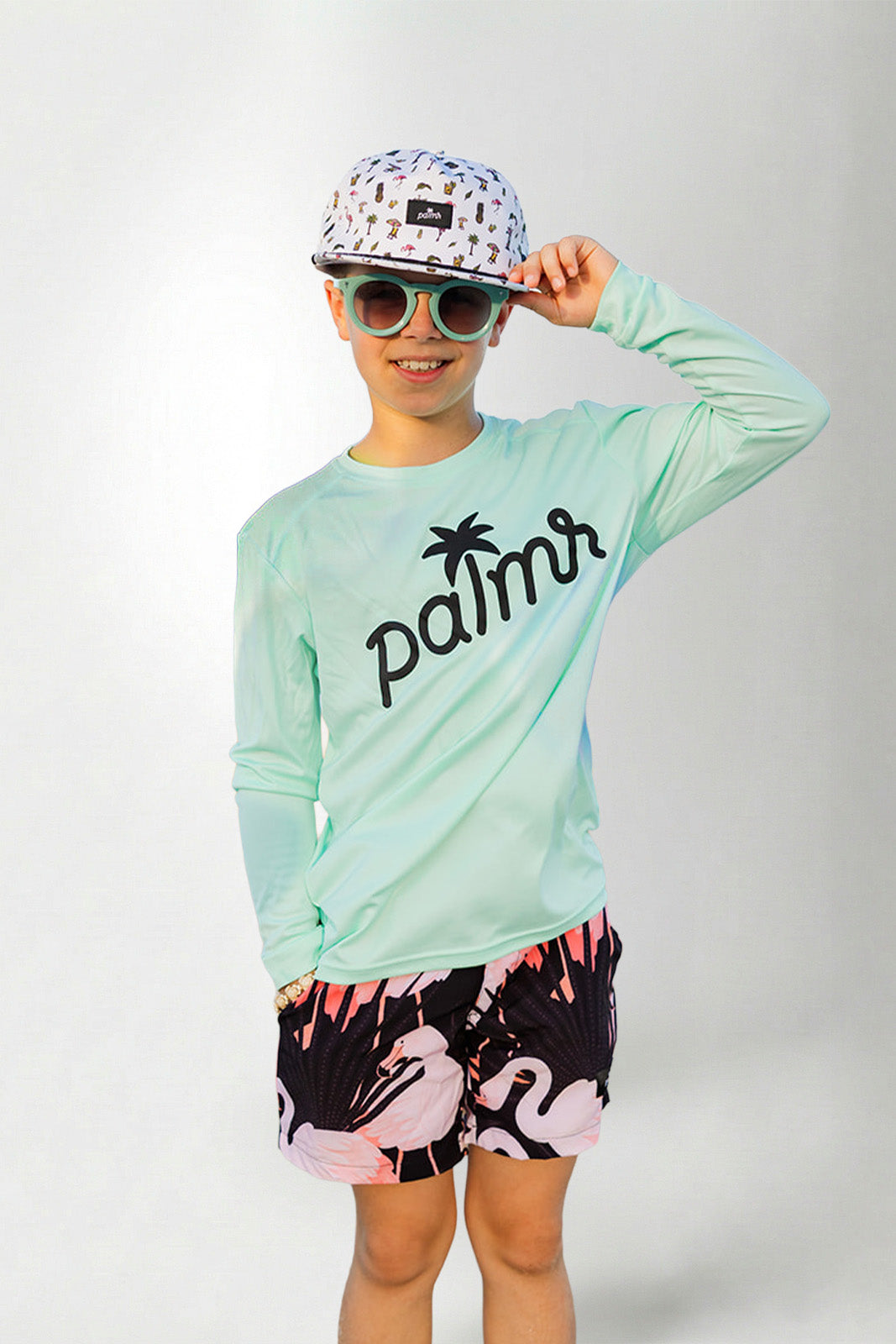 Person wearing a light green long-sleeve shirt with 'palms' logo, colorful cap, sunglasses, and flamingo shorts on a white background.