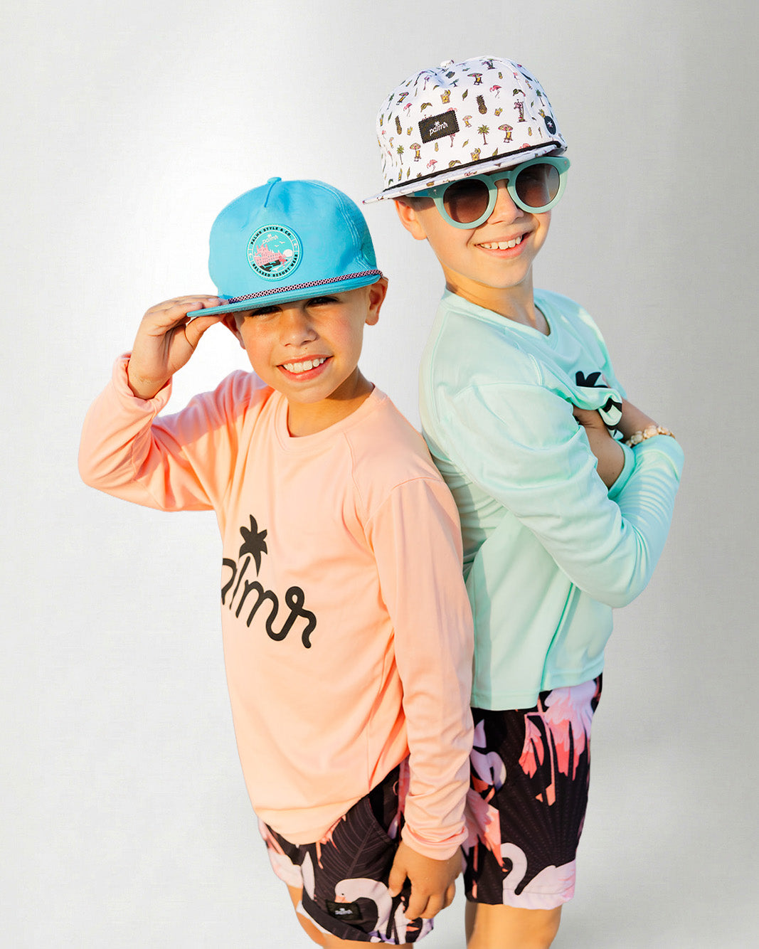 Person wearing a light green long-sleeve shirt with 'palms' logo, colorful cap, sunglasses, and flamingo shorts on a white background.