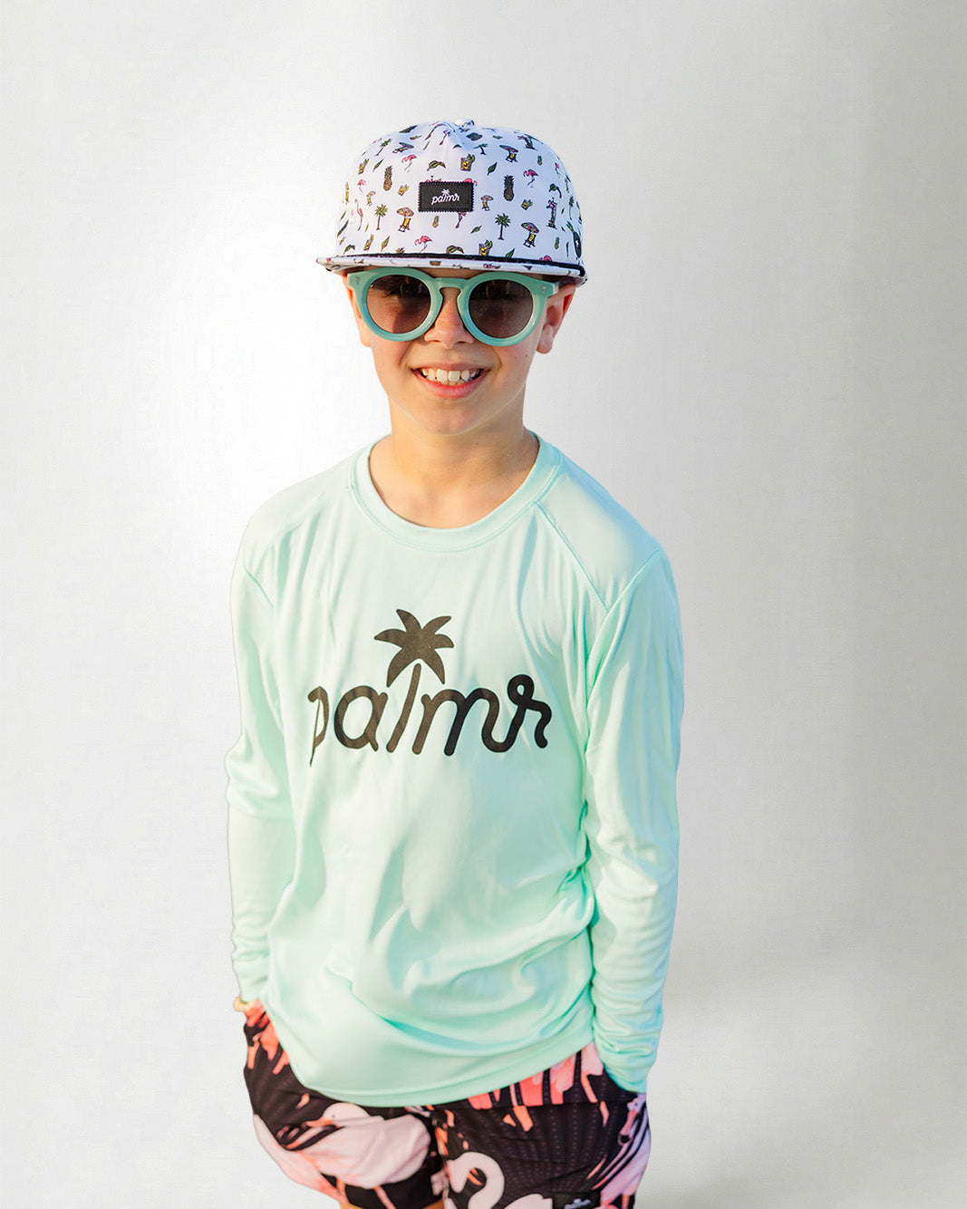 Person wearing a light green long-sleeve shirt with 'palms' logo, colorful cap, sunglasses, and flamingo shorts on a white background.