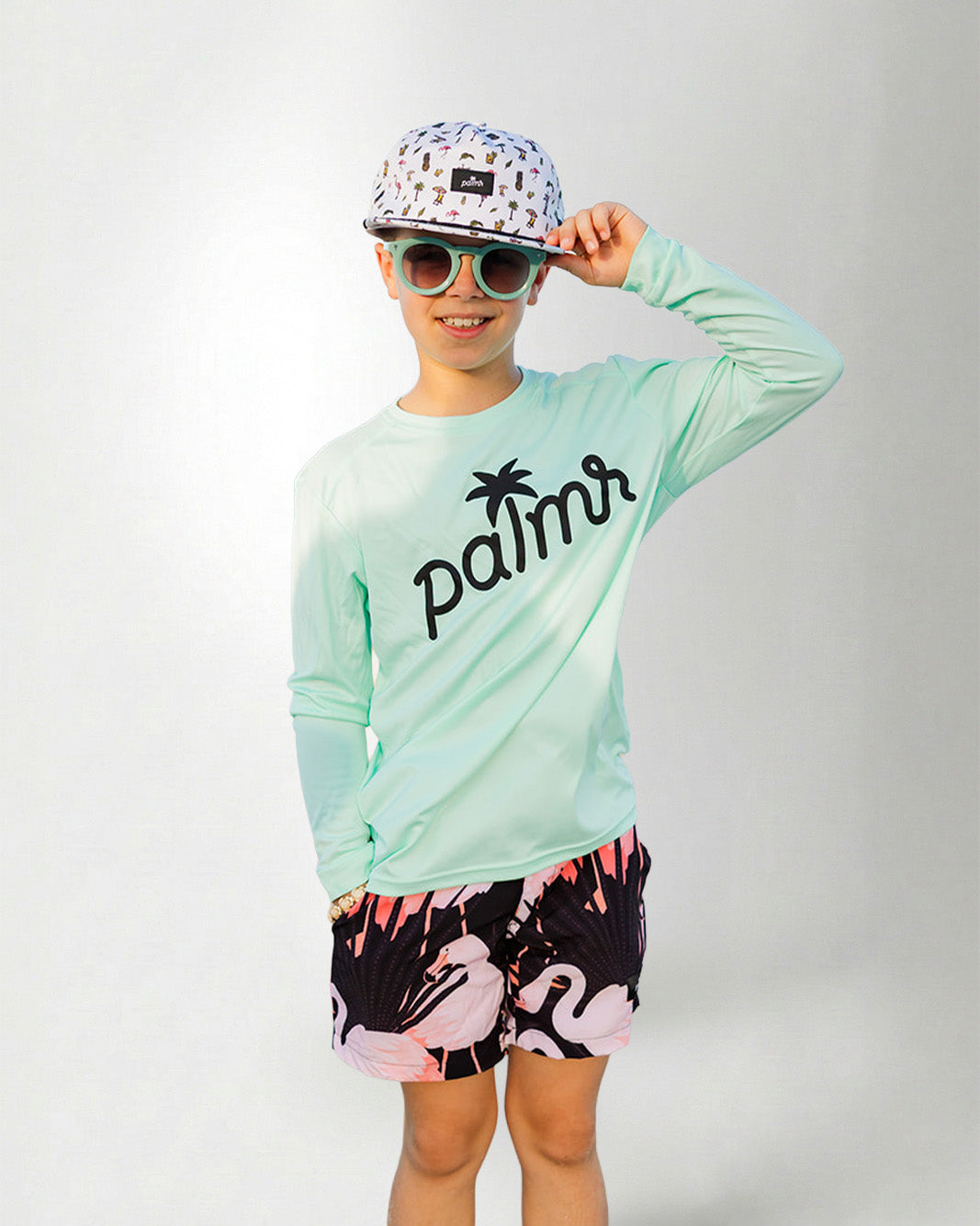 Person wearing a light green long-sleeve shirt with 'palms' logo, colorful cap, sunglasses, and flamingo shorts on a white background.