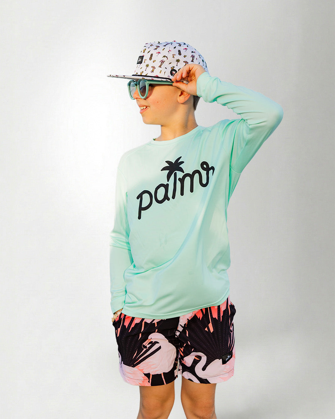 Person wearing a light green long-sleeve shirt with 'palms' logo, colorful cap, sunglasses, and flamingo shorts on a white background.