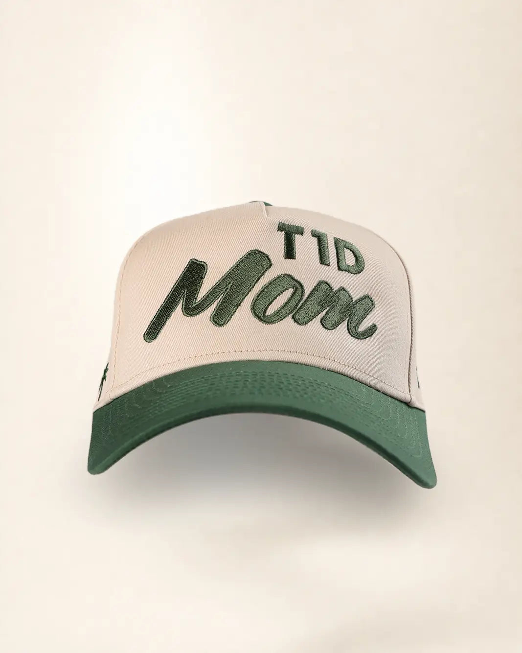 A structured trucker-style cap with a cream-colored front panel and brim, featuring dark green embroidered text reading 'T1D Mom' on the front, set against a plain light background.