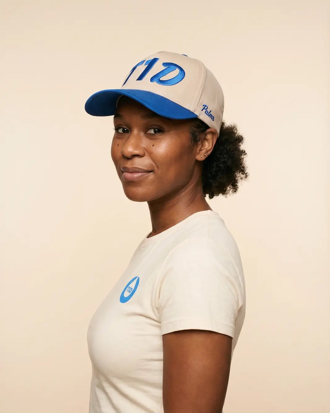 A woman wearing a cream-colored t-shirt and a matching cream and blue baseball cap, photographed against a solid light beige background.
